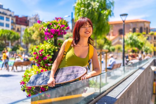 Smiling young chinese woman with skateboard gazing views form viewpoint in an urban square