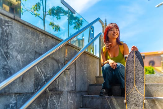 Cool asian young woman holding a skateboard sitting on stairs in the city in summer
