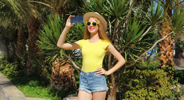 Summer vacation, happy smiling young woman taking selfie with phone in tourist straw hat, yellow glasses in the park with palm trees
