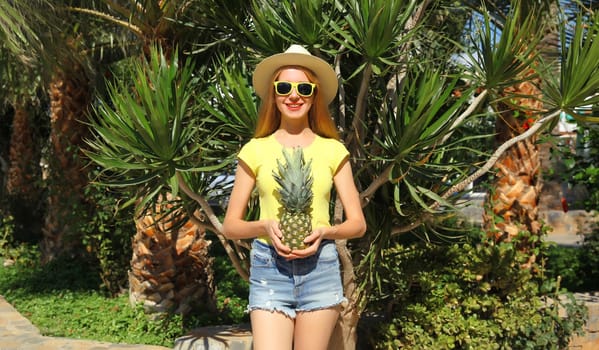 Summer vacation, tourism, happy smiling young woman with fruits, pineapple, joyful girl in glasses, straw tourist hat posing on the beach with palm tree
