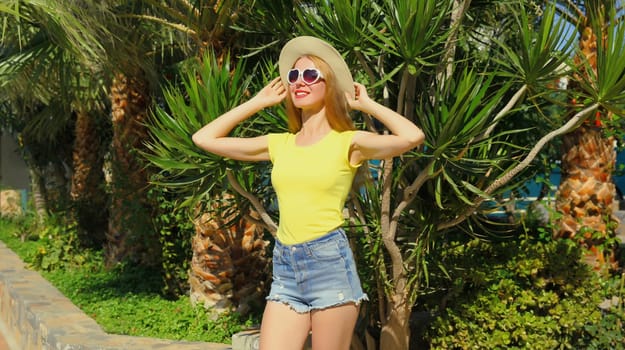 Summer vacation, happy beautiful young woman posing in tourist hat, glasses, yellow t-shirt, shorts, standing in park with palm trees