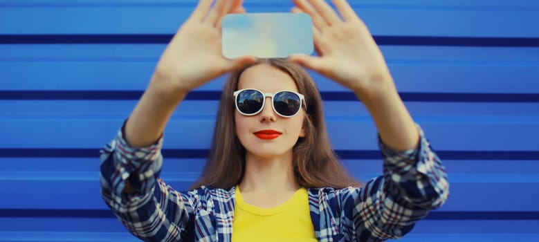 Close up of modern young woman taking selfie with phone, stylish cool girl using smartphone on blue wall background