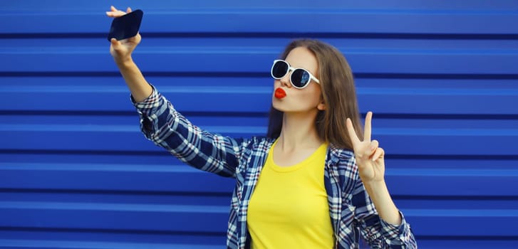 Summer cheerful stylish young woman taking selfie with smartphone, cool modern girl blowing a kiss in glasses on blue background