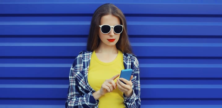 Stylish young woman with phone, modern girl looking at smartphone in casual clothing, glasses on blue wall background