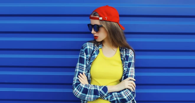 Portrait of stylish young girl in red baseball cap, glasses, modern teenager girl looking away posing on blue wall background