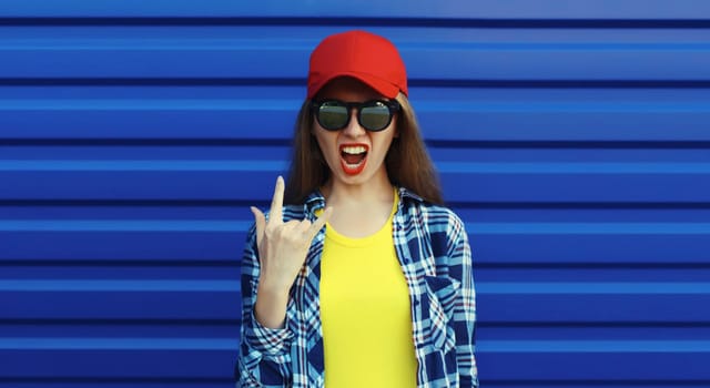 Stylish crazy young woman in glasses, cool modern teenage girl in red baseball cap posing on blue wall background
