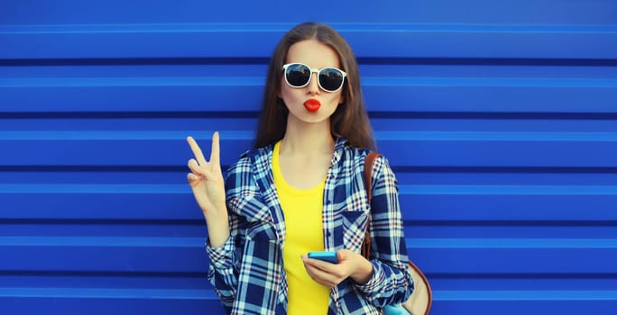 Portrait of stylish young woman blowing a kiss, cool modern girl posing with phone in casual clothing on blue wall background