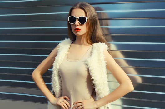 Portrait of modern stylish young woman in white fur vest, glasses, fashionable female model posing on gray silver wall background