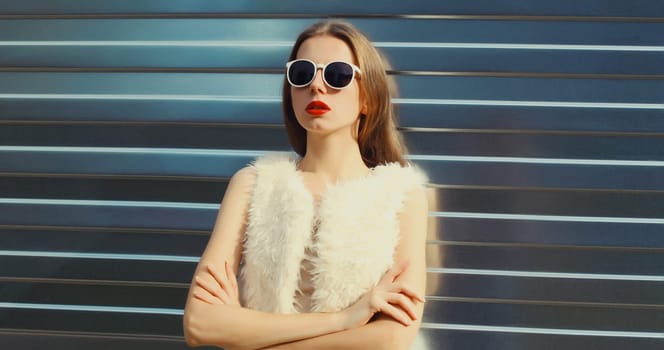 Portrait of modern stylish young woman in white fur vest, glasses, fashionable female model posing on gray silver wall background