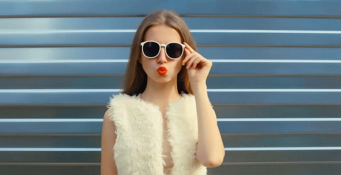 Portrait of modern stylish young woman blowing a kiss in white fur vest, glasses, fashionable female model posing on gray silver wall background