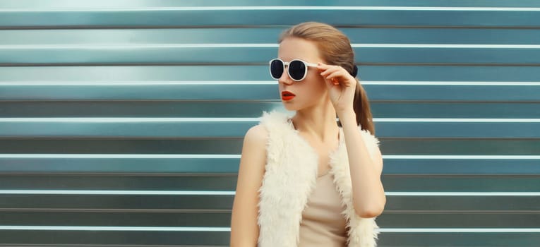 Portrait of modern stylish young woman in white fur vest, glasses, fashionable female model posing on gray silver wall background