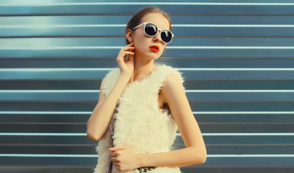 Portrait of modern stylish young woman in white fur vest, glasses, fashionable female model posing on gray silver wall background