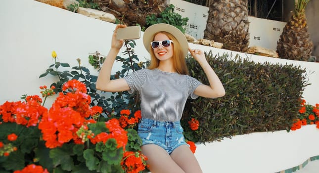 Summer vacation, happy smiling young woman taking selfie with phone, cheerful girl in tourist hat, glasses sitting in the park with red flowers