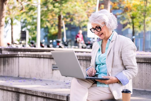 stylish mature woman smiling happy using laptop sitting outdoors in the city, concept of technology and urban lifestyle in adulthood, copyspace for text