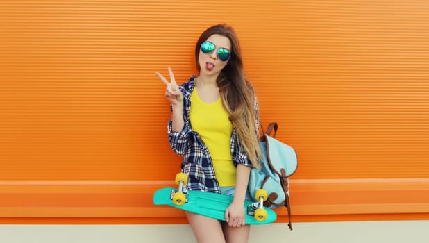 Summer portrait of happy cheerful stylish young woman with skateboard, teenager girl in colorful clothes, glasses posing on orange background on city street