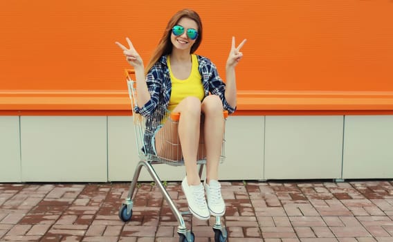 Stylish happy cheerful young woman, modern teenage girl smiling having fun sitting in shopping trolley cart on colorful orange background on city street