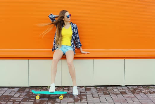 Summer image of stylish young woman with skateboard in colorful clothes looking away, modern teenage girl posing on orange background, blank copy space