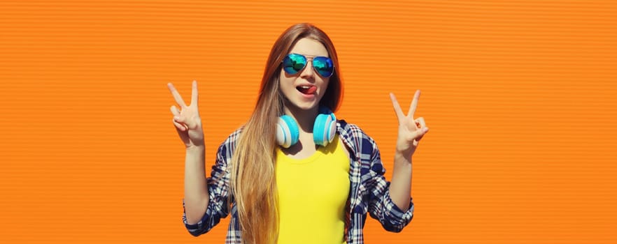 Stylish young woman listening to music with headphones, cool modern teenager girl posing on colorful bright orange background, summer day