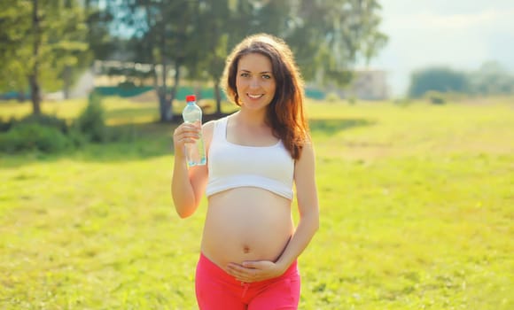 Pregnancy, maternity, healthy lifestyle, happy young pregnant woman drinking water from plastic bottle standing on the grass in sunny summer park