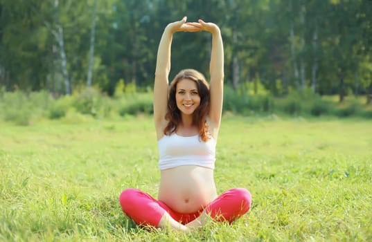 Pregnancy, fitness, happy young pregnant woman doing stretching exercises sitting on the grass in summer park