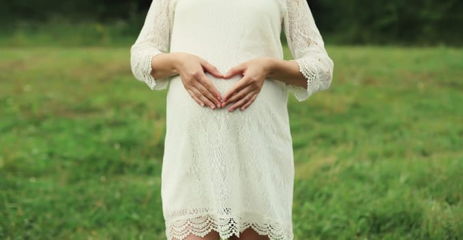 Pregnancy, maternity, happy pregnant woman makes heart sign on her belly with her hands in summer park