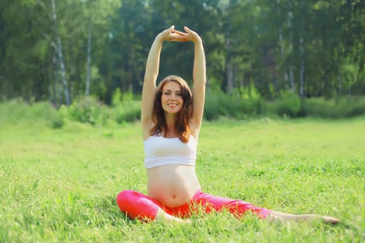 Pregnancy, fitness, happy young pregnant woman doing stretching exercises sitting on the grass in summer park