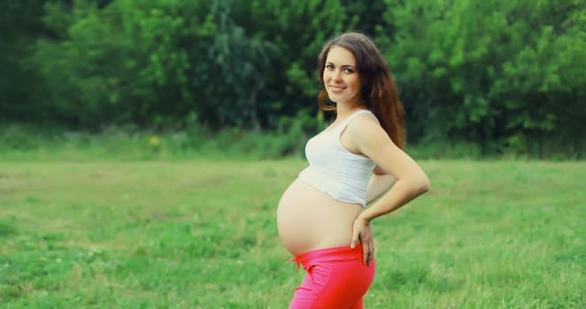 Portrait of beautiful happy smiling young pregnant woman looking at camera standing in summer park