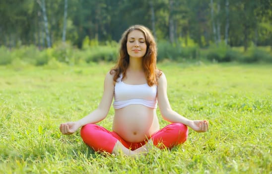 Pregnancy, fitness, happy young pregnant woman doing exercises sitting in lotus position on the grass in summer park