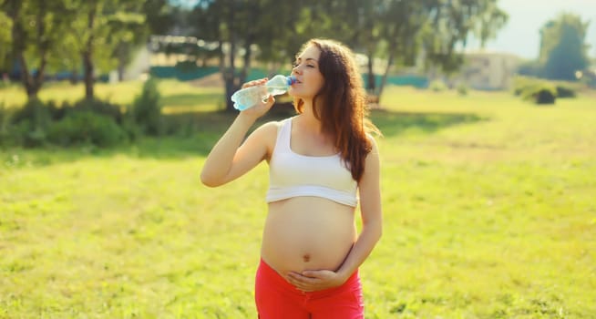 Pregnancy, maternity, healthy lifestyle, happy young pregnant woman drinking water from plastic bottle standing on the grass in sunny summer park