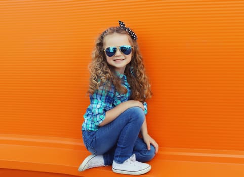 Happy cheerful kid girl with curly hair, joyful little child having fun on city street on orange wall background