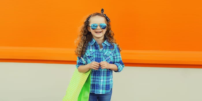 Stylish happy joyful kid girl standing with shopping bag, cheerful little girl child on city street on orange background