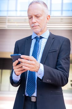 Satisfied with the results, the businessman walks down the street outside the office building, a mature boss holds a phone and coffee in his hands, writes messages and reads news online, using an app.