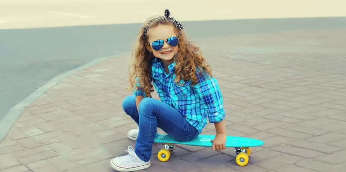 Happy joyful kid girl sitting on a skateboard, cheerful smiling little child on city street on a summer day