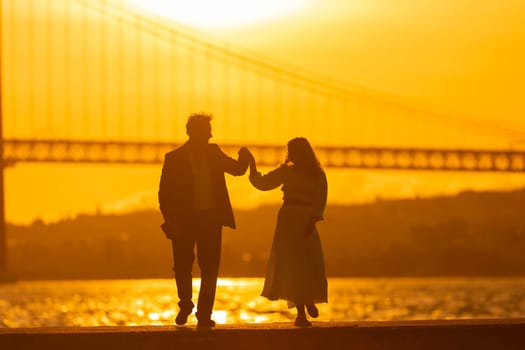 A couple dances in silhouette against a vibrant sunset with a bridge in the background.