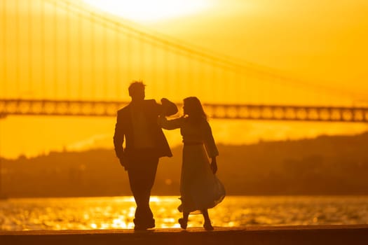 A man and woman dance in silhouette against a sunset with a bridge in the background.
