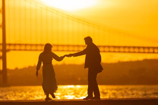 A man and woman dance in silhouette against a vibrant sunset, with a bridge in the background.