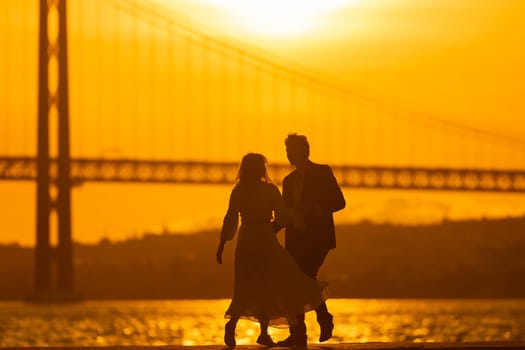 A couple dances together on a pier with a bridge in the background, silhouetted against the setting sun.