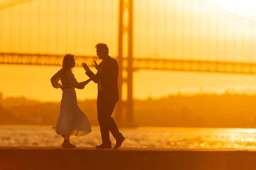 A silhouette of a couple dancing on a pier as the sun sets behind a bridge. The golden light of the sunset creates a romantic atmosphere.