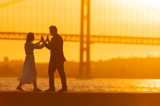 A silhouetted couple dances together on a waterfront as the sun sets behind a bridge.