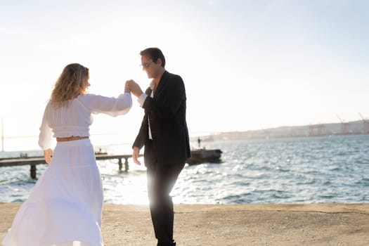 A woman in a white dress and a man in a suit dance together on a pier overlooking the ocean as the sun sets.
