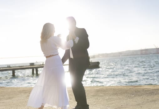 A woman in a white dress and a man in a suit dance by the ocean as the sun sets in the distance.