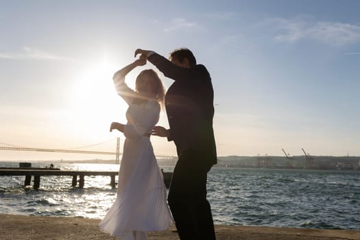 A man and woman dance on a waterfront pier as the sun sets behind them, casting a golden glow over the water.