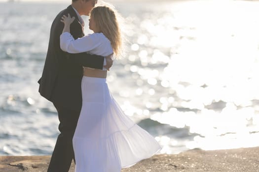 A couple dances on a rocky shore as the sun sets over the ocean. The woman wears a white dress and the man wears a suit.