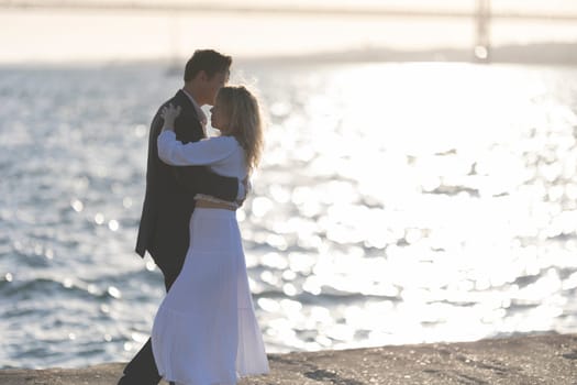 A couple dances on a rocky shore as the sun sets behind them, creating a warm glow over the water.
