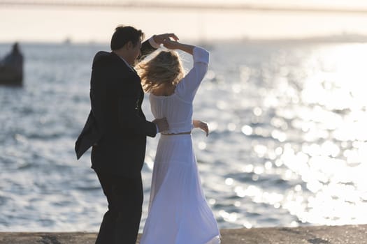 A woman in a white dress dances with a man in a suit, silhouetted against a golden sunset over the ocean.