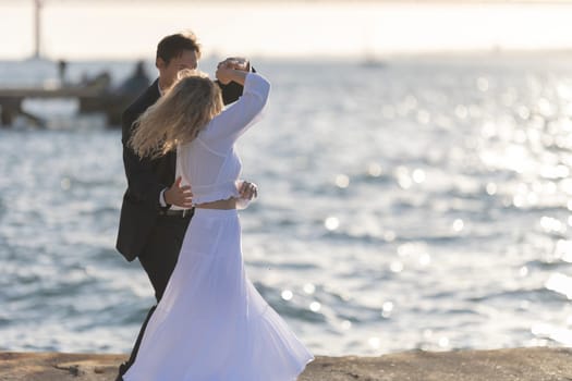 A woman and a man dance on a pier overlooking a sparkling sea at sunset. The woman wears a flowing white dress, and the man wears a dark suit.