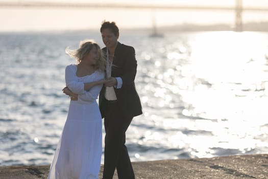 A couple dances by the ocean at sunset, the woman in a white dress and the man in a suit.