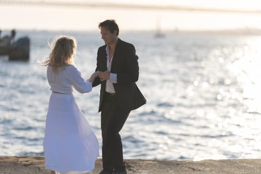A woman in a white dress dances with a man in a suit, smiling as the sun sets behind them and casts a golden glow on the ocean.