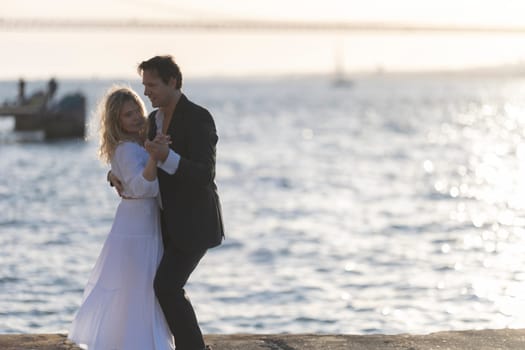 A couple dances on a pier as the sun sets over the water. They are silhouetted against the bright horizon, and their happy expressions are visible.