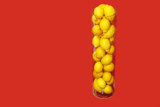 A glass transparent vase filled with fresh, ripe, yellow lemons stands on a red fabric background.
